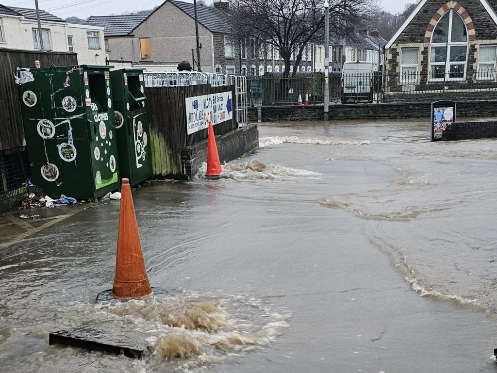 Urban street in Briton Ferry with water surging from a manhole, cones placed nearby.