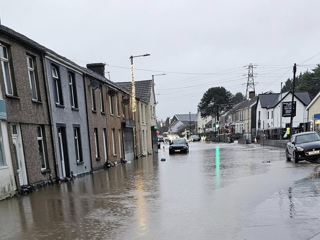 Flooded residential street in Briton Ferry with cars partially submerged in rising water.