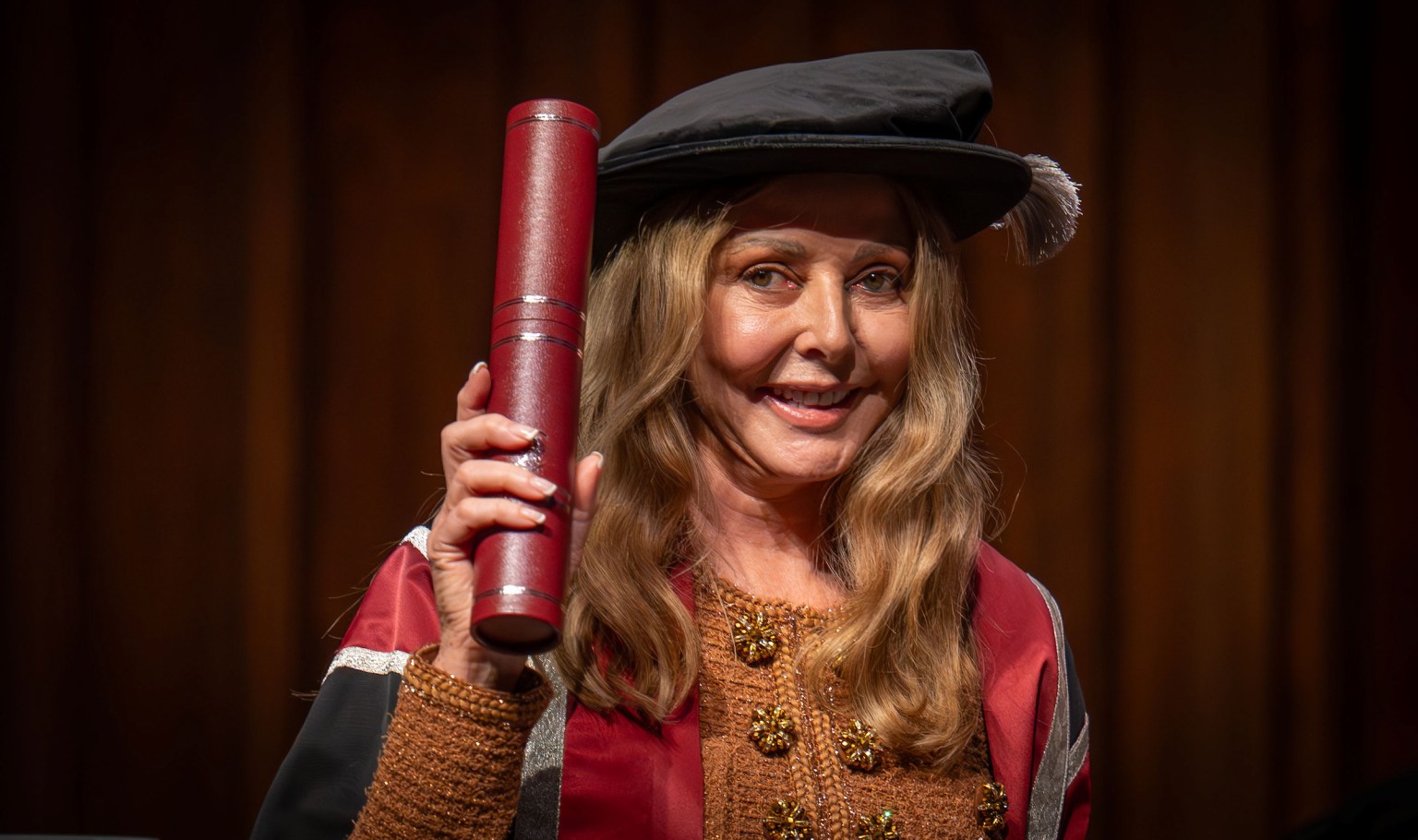 Carol Vorderman wearing academic gown and cap, holding a red diploma scroll at Swansea University graduation ceremony.