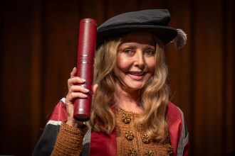 Carol Vorderman wearing academic gown and cap, holding a red diploma scroll at Swansea University graduation ceremony.