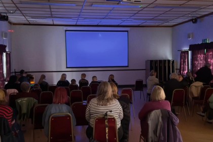 Audience of adults and children seated in Coelbren Welfare Hall facing a blank projection screen before a film screening.