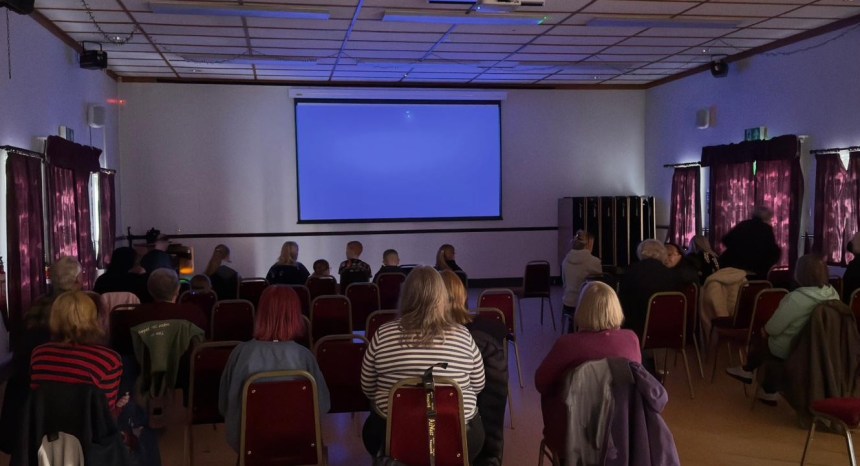 Audience of adults and children seated in Coelbren Welfare Hall facing a blank projection screen before a film screening.