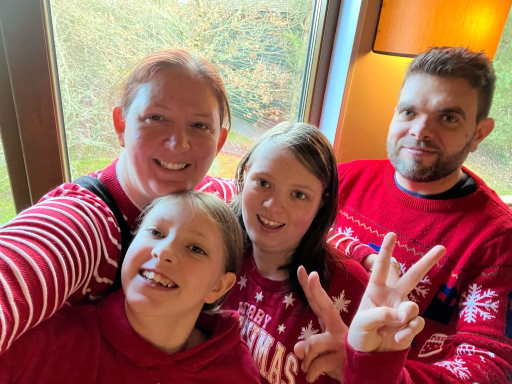 Four people in red Christmas sweaters smiling for a selfie by a window.