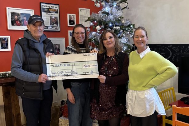 Four people standing indoors in front of a decorated Christmas tree, holding a large ceremonial cheque for £600. The setting includes festive decorations, framed photos, and a chalkboard wall with drawings and text.