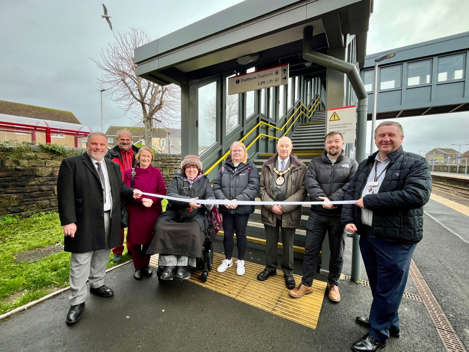 Nine people, including one in a wheelchair, stand in front of a new lift entrance at Llanelli Station during a ribbon-cutting event.