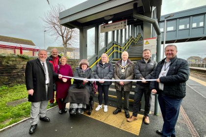 Nine people, including one in a wheelchair, stand in front of a new lift entrance at Llanelli Station during a ribbon-cutting event.