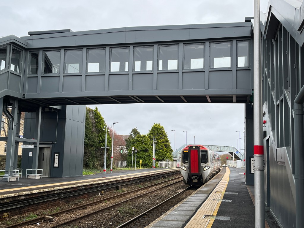 Railway station with a glass-panelled overpass and visible elevator shaft, designed for accessible platform crossing.