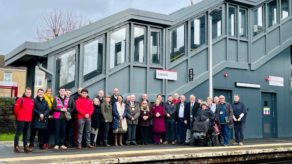 Group of people pose on Llanelli Station platform in front of a modern lift and stairwell structure, with station signage visible.