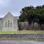 Exterior of St David’s Church in Penllergaer, showing grey stone walls, Gothic windows, and mature trees under an overcast sky.