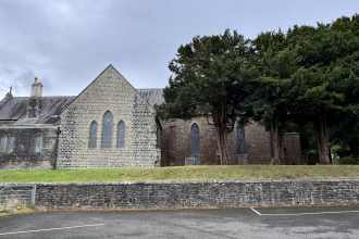 Exterior of St David’s Church in Penllergaer, showing grey stone walls, Gothic windows, and mature trees under an overcast sky.
