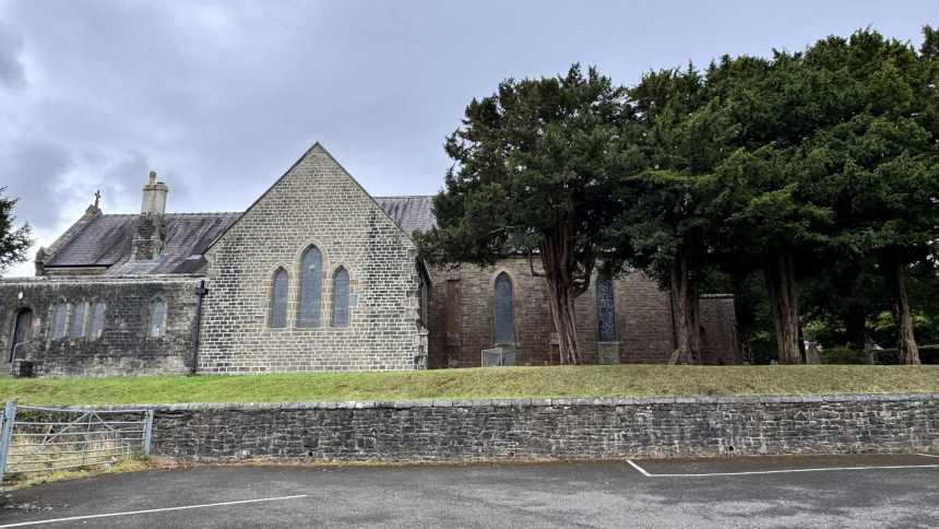 Exterior of St David’s Church in Penllergaer, showing grey stone walls, Gothic windows, and mature trees under an overcast sky.