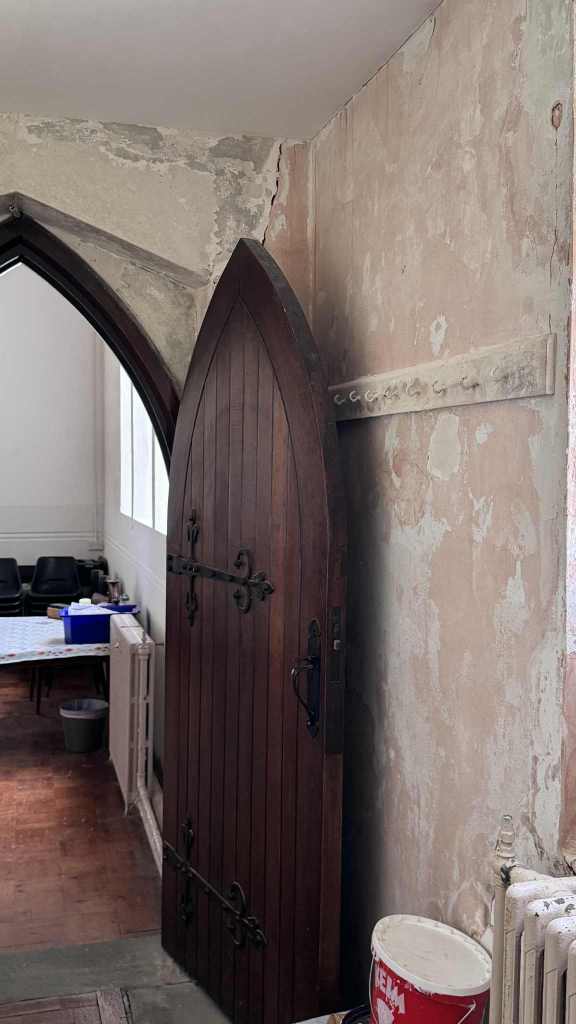 Gothic-style wooden door in St David’s Church with ornate hinges, surrounded by cracked plaster and a paint bucket.