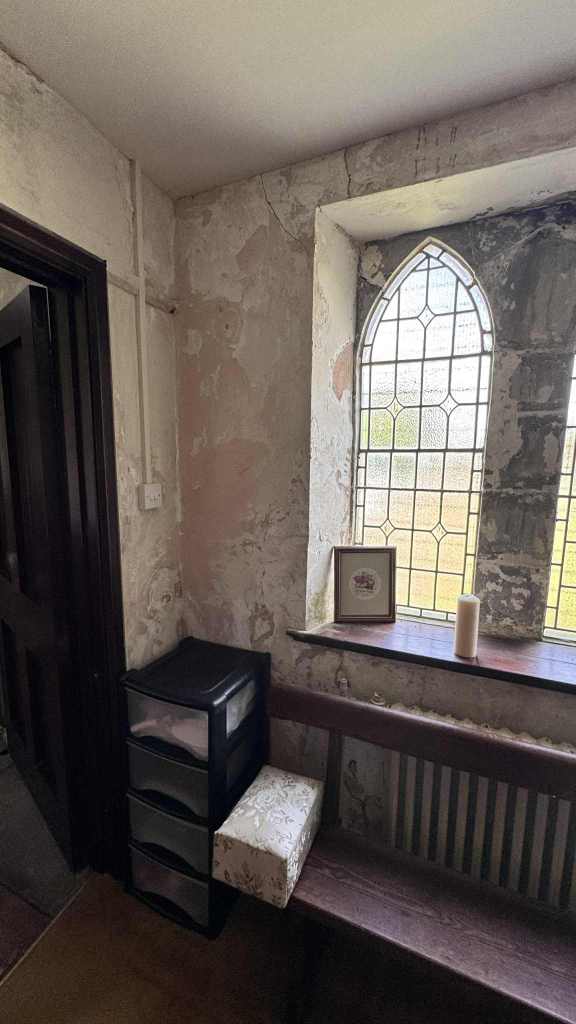 Bench and candle beneath a tall arched window in St David’s Church, with peeling plaster and rustic furnishings.