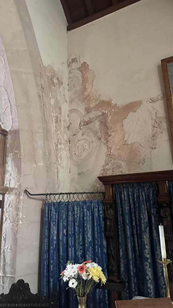 Peeling plaster and water damage on an interior wall of St David’s Church, with brass candle holder and floral arrangement below.