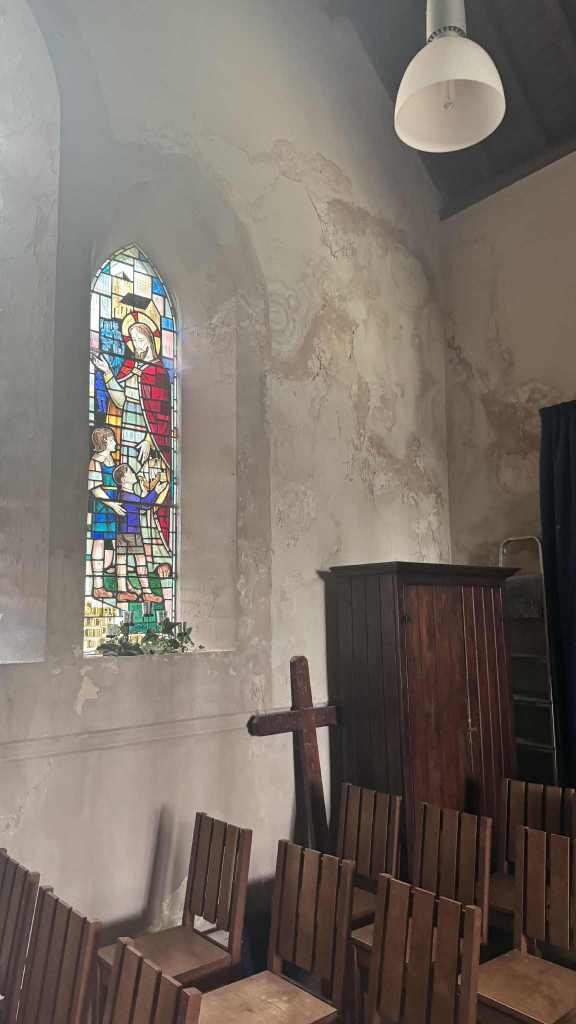Stained glass window in St David’s Church depicting a religious figure with two children, surrounded by aged walls and wooden chairs.