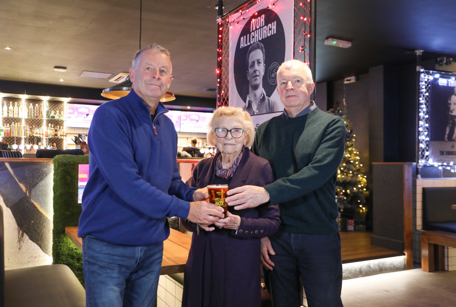 Esme Allchurch stands with family at The Swansea Jack sports bar, holding a pint of Golden Boy lager beside a tribute to Swansea City legend Ivor Allchurch.