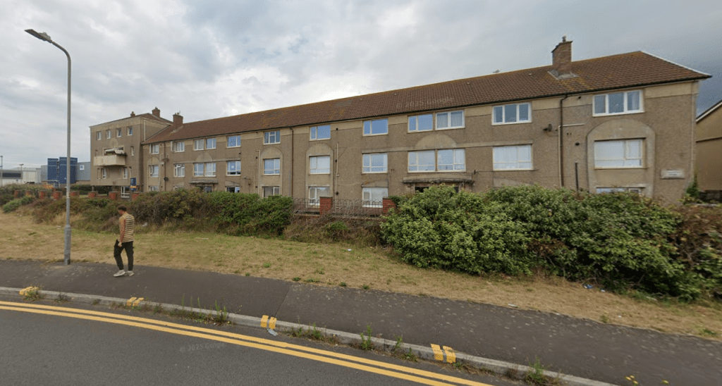 Three-storey residential block with tiled roof and beige walls, viewed from roadside with grassy verge and footpath.
