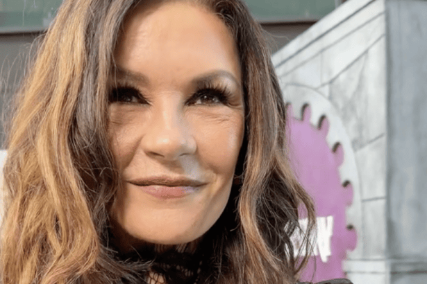 Catherine Zeta‑Jones speaking during a TikTok interview at a Netflix red carpet event. She is wearing a black lace top and smiling slightly, with a purple gear‑shaped backdrop behind her. The setting is outdoors in an urban environment.