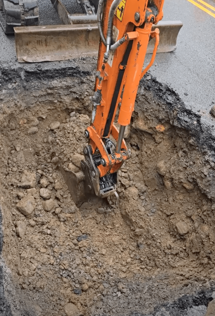 Excavator digging into broken asphalt at Brynymor Road sinkhole site.