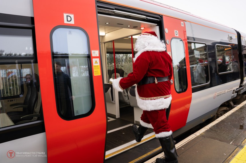 Person dressed as Santa Claus stepping onto a Transport for Wales train at a station, with festive reflections in the windows.