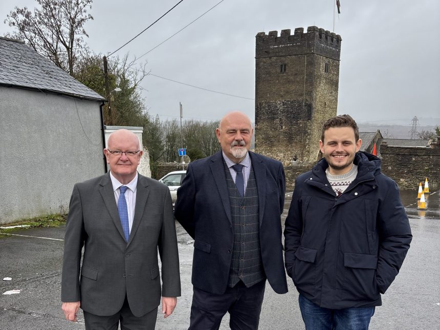 Three men stand in front of the crenellated stone tower of St David & St Cyfelach’s Church in Llangyfelach. From left to right are Cllr Lyndon Jones, Cllr Mark Tribe and Tom Giffard MS. They are standing on a wet pavement with the historic church tower behind them.