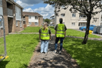Two Carmarthenshire County Council officers in high-visibility vests stand on a pathway in a residential area, facing a group of apartment buildings during a community inspection.