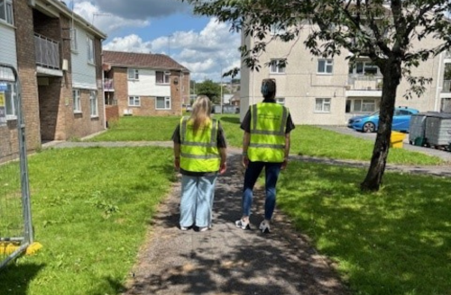 Two Carmarthenshire County Council officers in high-visibility vests stand on a pathway in a residential area, facing a group of apartment buildings during a community inspection.