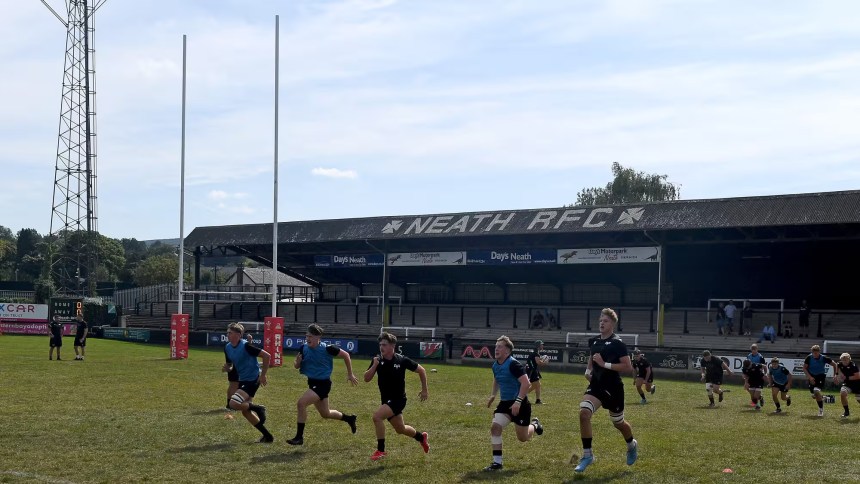 Young rugby players in black and blue kit sprinting in formation on the pitch at Neath RFC’s home ground, The Gnoll, during a training session.