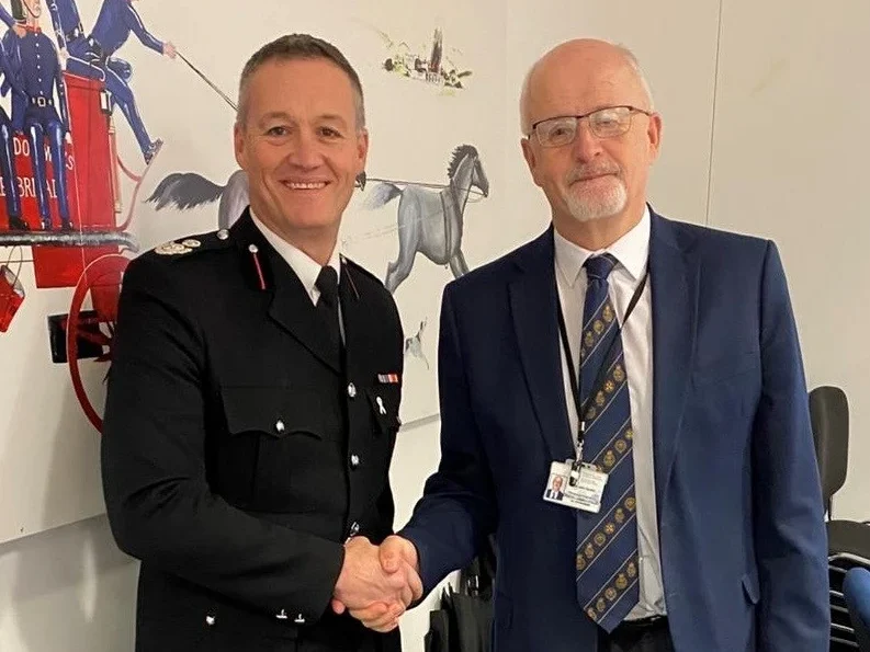 Craig Flannery in uniform shaking hands with Cllr John Davies in front of a fire service mural.