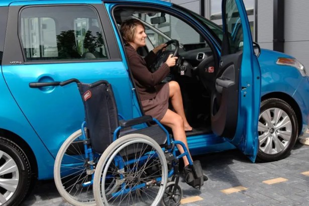 A person seated in a blue car with the driver’s door open, next to a wheelchair on a paved area.