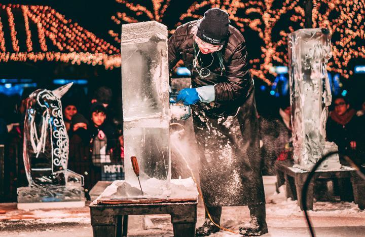 Person carving ice sculpture at outdoor winter festival in Swansea, with string lights and crowd watching.
