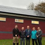 Five people stand in front of a brick building with solar panels installed on the roof. The building has red and blue trim and four windows. The group is posing outdoors on grass, with trees in the background, highlighting the club’s new renewable energy setup.