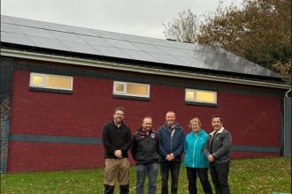 Five people stand in front of a brick building with solar panels installed on the roof. The building has red and blue trim and four windows. The group is posing outdoors on grass, with trees in the background, highlighting the club’s new renewable energy setup.