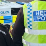 Two South Wales Police officers in uniform, one wearing a high-visibility vest, standing near a marked police van.