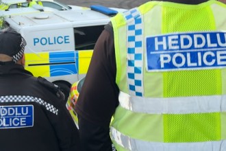 Two South Wales Police officers in uniform, one wearing a high-visibility vest, standing near a marked police van.