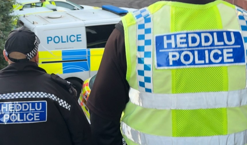 Two South Wales Police officers in uniform, one wearing a high-visibility vest, standing near a marked police van.