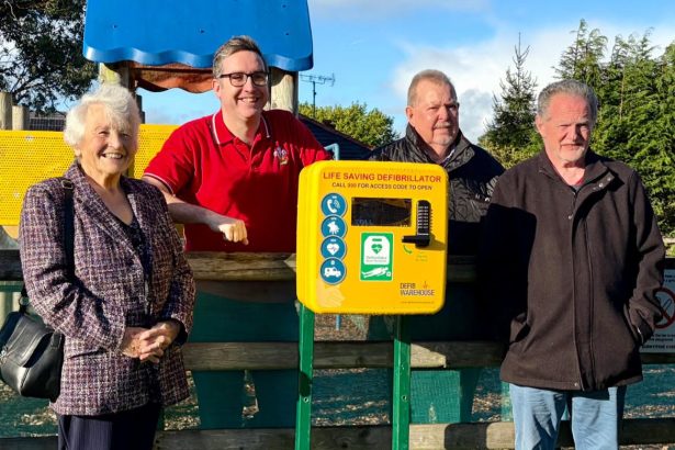 Four Llanelli town councillors stand beside a yellow defibrillator unit at Nightingale Court play area in Bigyn.