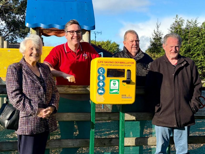 Four Llanelli town councillors stand beside a yellow defibrillator unit at Nightingale Court play area in Bigyn.