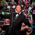 Sir Bryn Terfel singing on stage mid‑performance, with an orchestra behind him and purple and white flowers arranged around the stage.