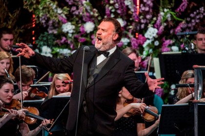 Sir Bryn Terfel singing on stage mid‑performance, with an orchestra behind him and purple and white flowers arranged around the stage.