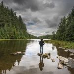 Person standing on rocks beside a forested lake under dramatic, overcast skies.