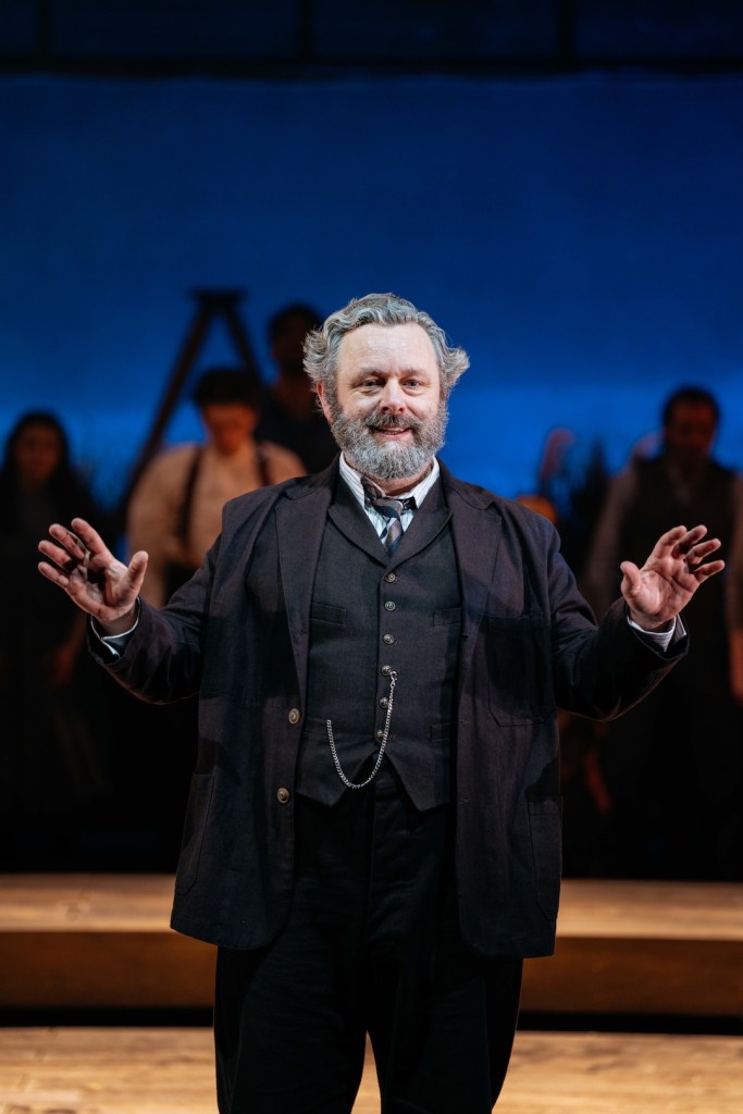 Michael Sheen in a spotlight delivering a monologue in Our Town, with ensemble behind him. Image: Welsh National Theatre.
