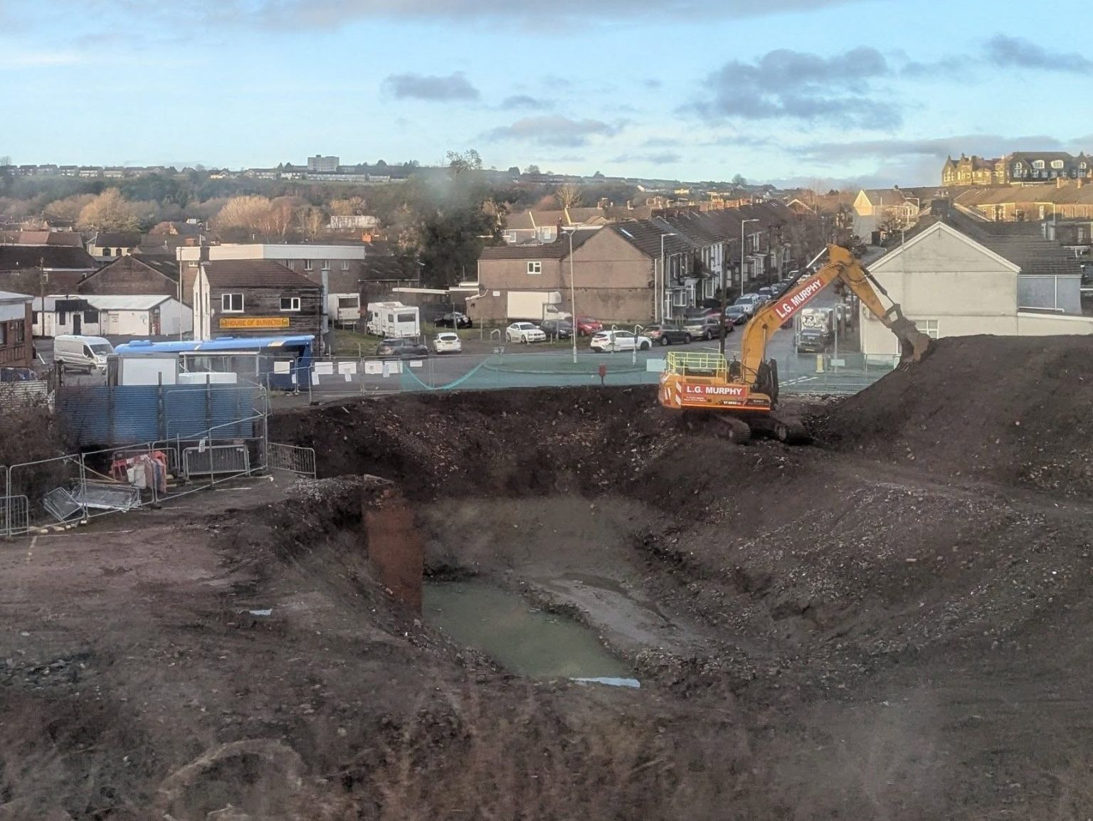Excavator digging a deep pit at the former Cwmfelin Social Club site in Swansea, with construction fencing and pumping equipment visible.