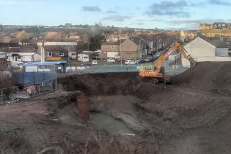 Excavator digging a deep pit at the former Cwmfelin Social Club site in Swansea, with construction fencing and pumping equipment visible.
