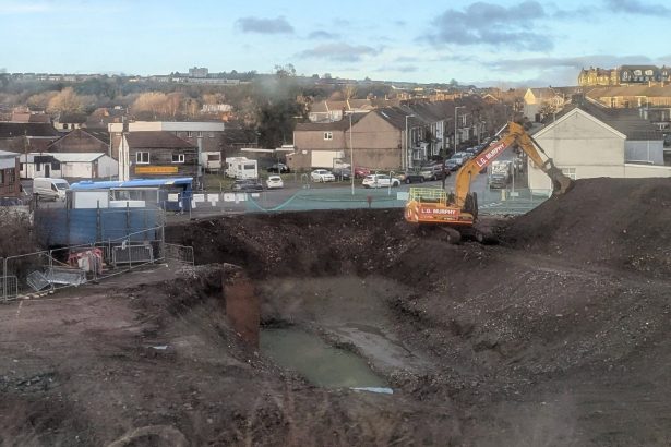 Excavator digging a deep pit at the former Cwmfelin Social Club site in Swansea, with construction fencing and pumping equipment visible.
