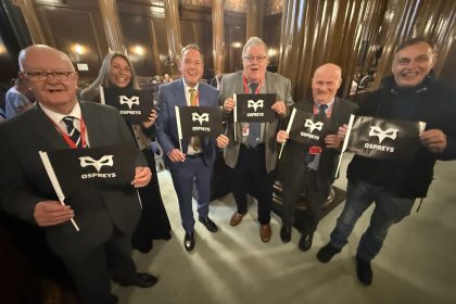 Group photo of Swansea councillors from all parties and the council’s chief executive in the Guildhall chamber holding Ospreys signs.