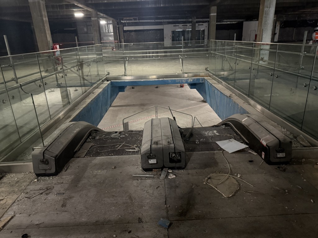 Top of escalators inside the old Debenhams, with exposed concrete floors and construction lighting.