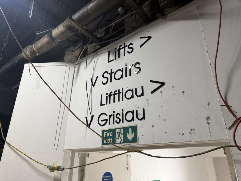 Bilingual directional sign for lifts and stairs inside empty Debenhams interior.