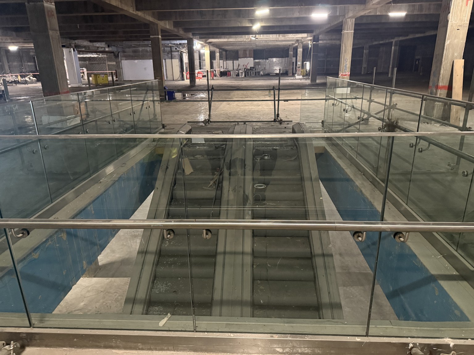 View down escalators inside empty Debenhams with exposed ceiling and construction fencing.