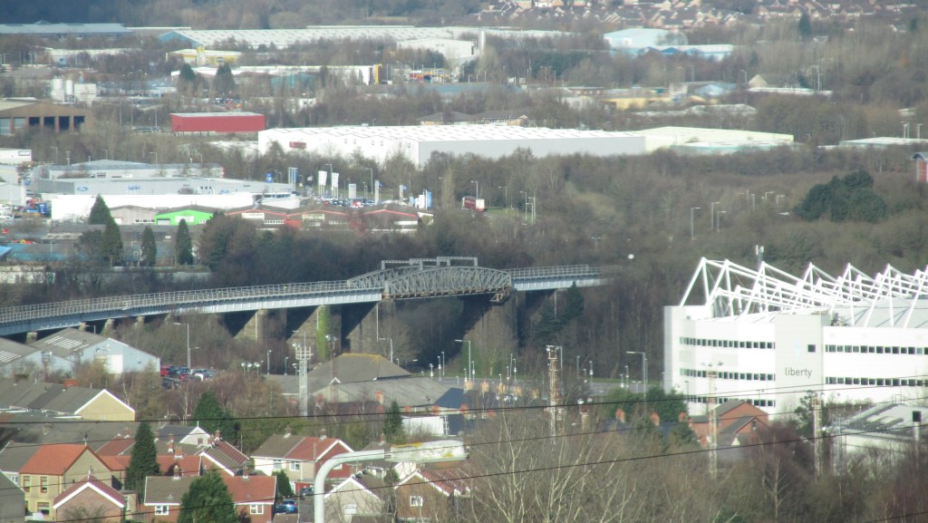 Steel truss railway viaduct supported by stone pillars near Swansea Stadium, surrounded by urban and industrial buildings.